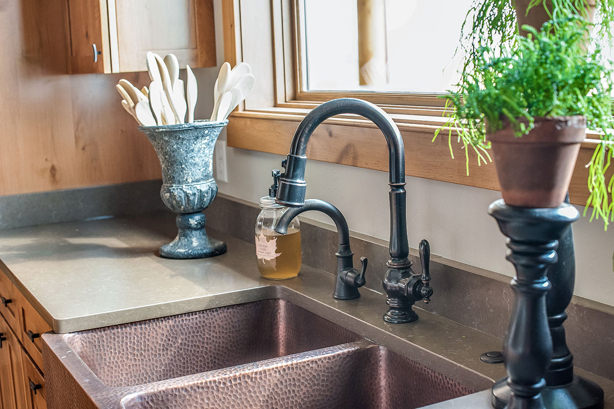 A kitchen counter with a vase full of utensils next to a copper double basin farmhouse sink with rubbed bronze fixtures
