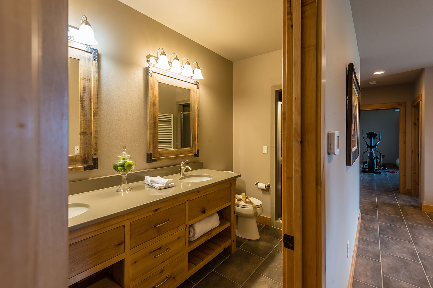 Peeking into bathroom with dual vanities next to a toilet with a bow on it and workout room in distance