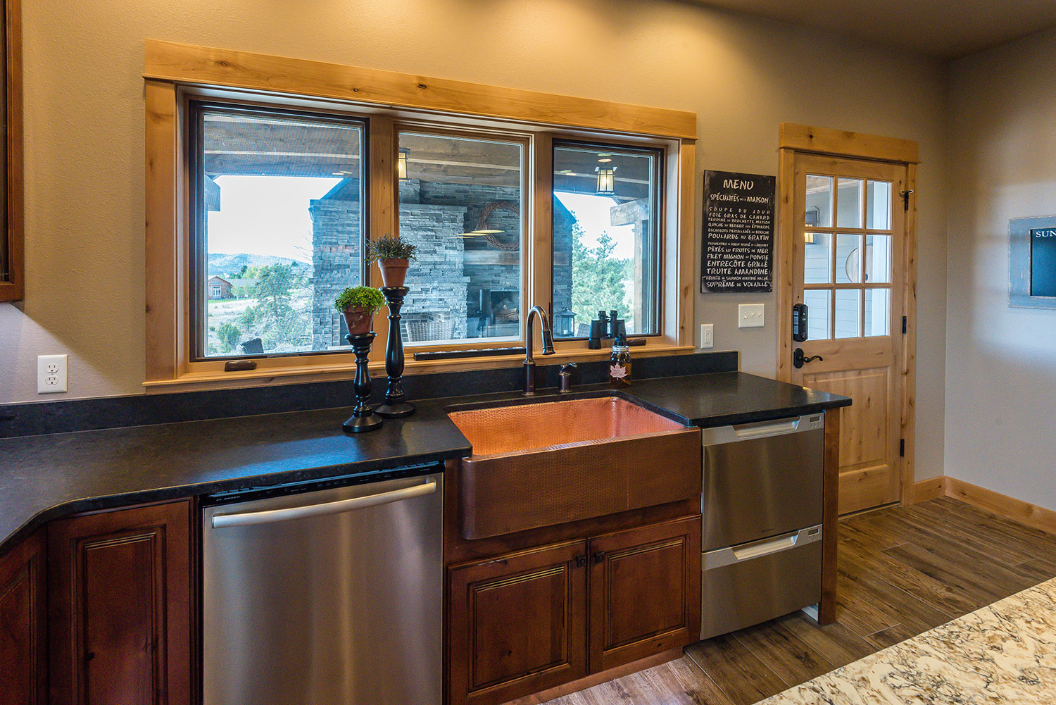 View of a copper farmhouse sink between a dishwasher on the left and an under counter refrigerator on the right below awindow