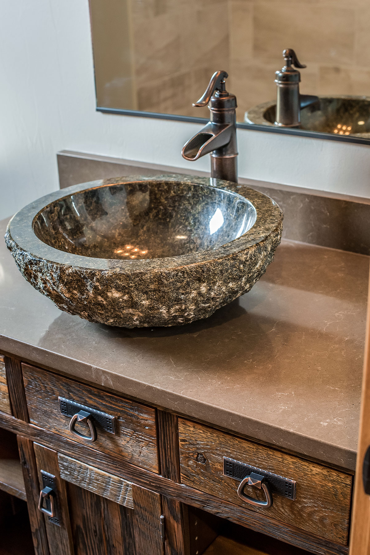 Vanity with rustic cabinets and granite countertop with a large carved rock basin sink and waterfall faucet in below a mirror