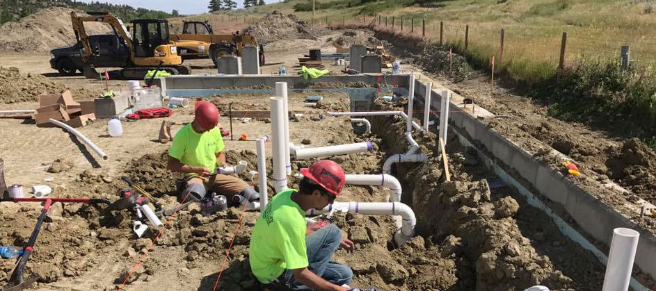 Workers in high visibility shirts and hard hats installing plumbing in a building under construction with heavy equipment